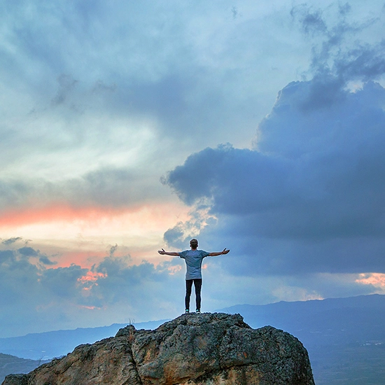 Un homme debout au sommet d'un rocher avec les bras ouverts face à un ciel nuageux au coucher du soleil.